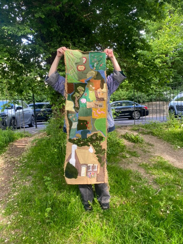 image of someone outside holding up a hand knitted tapestry depicting patchwork farmland